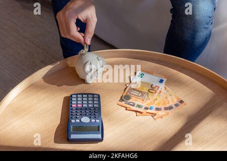 Personne méconnaissable insérant une pièce d'euro dans une banque de porc comme concept d'économie d'argent avec une calculatrice et des factures à la maison dans le salon Banque D'Images