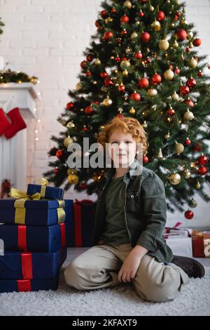 un garçon redhead assis sur le sol près des cadeaux de Noël et un pin décoré de boules à la maison Banque D'Images