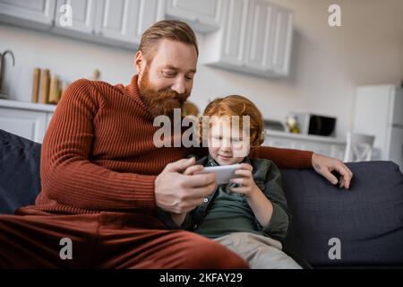 homme barbu souriant assis sur un canapé près d'un fils roux à l'aide d'un téléphone portable Banque D'Images