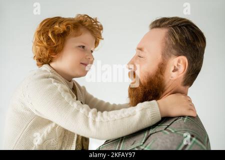 Sourire rouge poil enfant embrassant le père barbu dans une veste isolée sur gris Banque D'Images