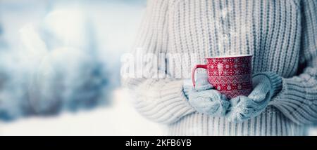 femme en laine chandail et gants mitaines tenant une tasse de boisson chaude à la vapeur sur fond de paysage hivernal neigeux. bannière avec espace de copie Banque D'Images