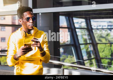 Portrait d'un jeune Africain fumant vêtu d'un sweat-shirt jaune et de lunettes de soleil tenant une cigarette avec une main et son téléphone portable avec l'OT Banque D'Images