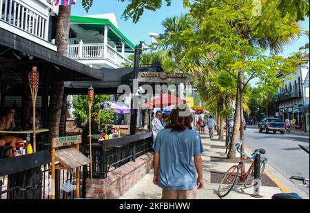 Long garçon aux cheveux marchant sur le trottoir à côté des restaurants et des arbres tropicaux avec des cyclistes et d'autres touristes à Key West Florida USA 21 juillet 2010 Banque D'Images