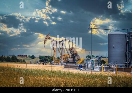 Deux crics de pompe et réservoirs et équipement de champ de pétrole dans la zone clôturée dans les pâturages sous un ciel spectaculaire avec des maisons sur la colline à l'horizon - été Banque D'Images