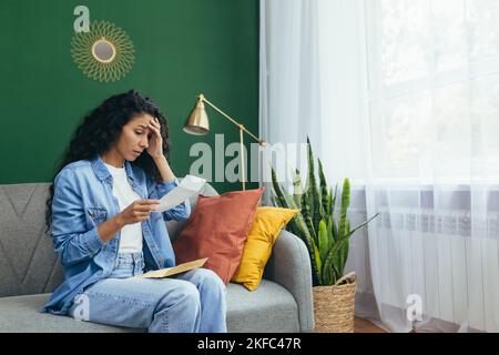 Une jeune femme latino-américaine a reçu des papiers de divorce. Assise à la maison sur le tapis, tenant une feuille de papier dans ses mains, choquée, tenant sa tête. Banque D'Images