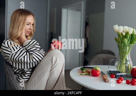 La jeune blonde enceinte dans les vêtements de la maison est assise à table dans sa cuisine et tient la pomme juteuse rouge dans ses mains. Banque D'Images