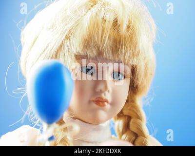 Poupée vintage fille blonde en porcelaine avec des tresses aux yeux bleus dans une robe légère avec des ballons blancs, roses et bleus. Des poupées en porcelaine sont apparues au 18th siècle en France. Ils ont été utilisés pour promouvoir la mode française dans le monde. Habituellement, de telles poupées étaient fournies avec un grand nombre de vêtements et envoyées aux clients sous forme de mini mannequins. Un peu plus tard, la production de poupées a été mise sur une base industrielle et les enfants de parents riches ont commencé à jouer avec eux. Naturellement, ces belles pièces ont attiré l'attention des collectionneurs et sont devenues encore plus populaires. En fait, la tête, les bras et les jambes sont m Banque D'Images