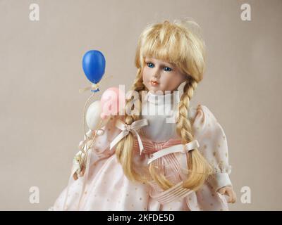 Poupée vintage fille blonde en porcelaine avec des tresses aux yeux bleus dans une robe légère avec des ballons blancs, roses et bleus. Des poupées en porcelaine sont apparues au 18th siècle en France. Ils ont été utilisés pour promouvoir la mode française dans le monde. Habituellement, de telles poupées étaient fournies avec un grand nombre de vêtements et envoyées aux clients sous forme de mini mannequins. Un peu plus tard, la production de poupées a été mise sur une base industrielle et les enfants de parents riches ont commencé à jouer avec eux. Naturellement, ces belles pièces ont attiré l'attention des collectionneurs et sont devenues encore plus populaires. En fait, la tête, les bras et les jambes sont m Banque D'Images