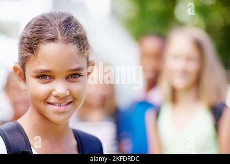 Je suis fier de mon école. Mignonne jeune fille multi-ethnique souriant à vous avec des camarades d'école flou en arrière-plan - espace d'imitation. Banque D'Images