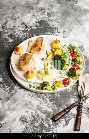 Filet de poisson de mer cuit avec des légumes. Filet de tilapia au brocoli, tomates cerises, pommes de terre frites, pois verts et laitue. Fruits de mer sur un plat blanc Banque D'Images