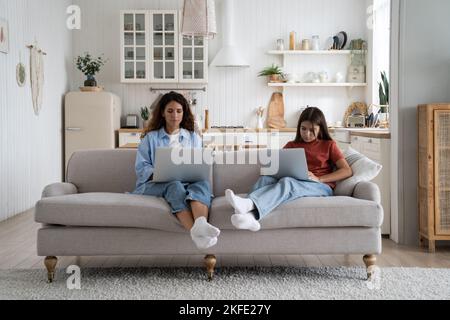 Femme insociable introvertie et adolescente en vêtements décontractés assis sur un canapé avec des ordinateurs portables sur des longueurs Banque D'Images