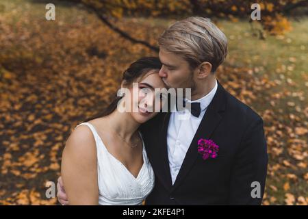 Belle mariée turque à sa fin 20s penchée la tête au-dessus du bras de son conjoint scandinave. Blonde barbu homme beau embrassant sa femme sur son front. Photo de mariage dans le parc d'automne recouvert de feuilles d'orange et d'or. Photo de haute qualité Banque D'Images