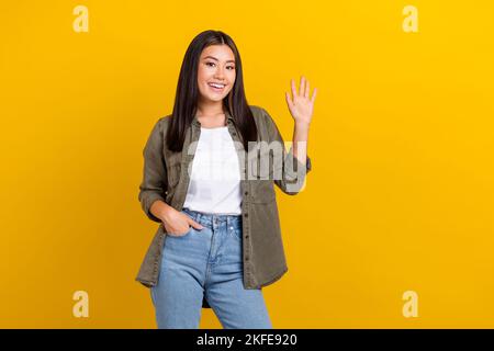 Portrait de bonne humeur satisfait belle fille longue coiffure porter chemise kaki tenir le bras dans la poche en agitant la main isolée sur fond jaune Banque D'Images