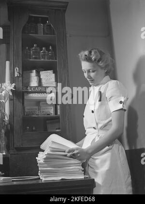 Un rasage sur le 1940s. Une jeune femme dans un salon de coiffure qui a l'air très belle et rangée avec elle dans une tenue de travail appropriée avec des serviettes fraîches et des produits de beauté de rasage et de cheveux visibles dans le placard. Suède mai 1940 Kristoffersson réf. 151 Banque D'Images