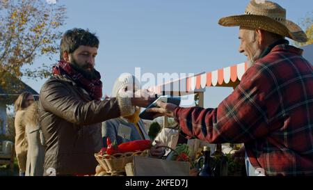 Les conjoints magasinent sur le marché agricole local. La femme se tient avec un panier de fruits ou de légumes. Le mari paie l'achat sans contact. Plats végétariens et bio. Agriculture. Banque D'Images