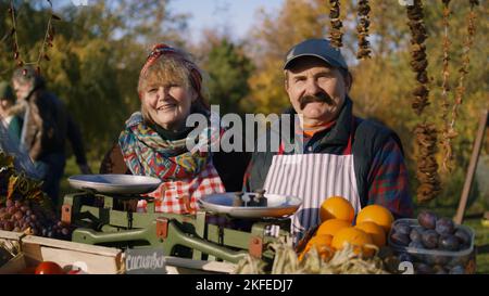 Heureux couple de personnes âgées d'agriculteurs se tenant près de fruits et légumes frais, regardant l'appareil photo, souriant. Faire du shopping sur le marché agricole local en plein air. Plats végétariens, biologiques et sains. Agriculture. Banque D'Images