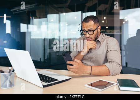 Pensant homme d'affaires afro-américain lisant des informations sur smartphone, homme travaillant au bureau au travail en utilisant un ordinateur portable, travailleur en chemise parcourant des pages en ligne sur téléphone sérieux et concentré. Banque D'Images