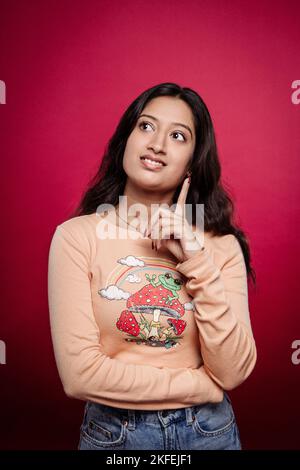 Jeune femme indienne souriante regardant sur le côté et pensant quelque chose en se tenant sur fond rouge. Une fille indienne fait des plans pour faire du shopping. Banque D'Images