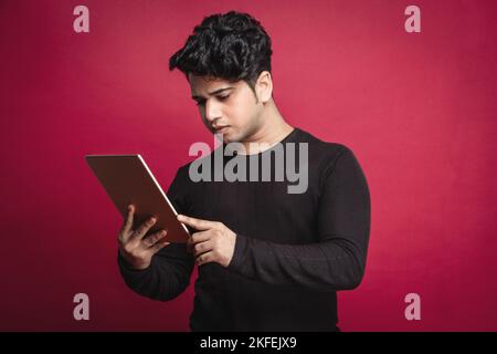 Jeune Indien sérieux dans un t-shirt noir utilisant une tablette numérique sur fond rouge Banque D'Images