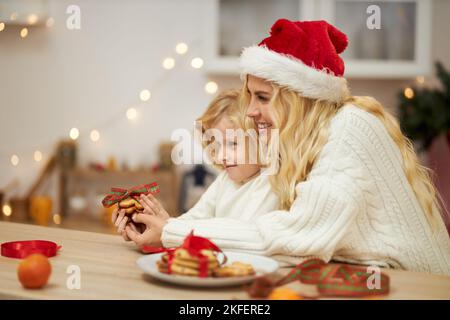 Vue latérale de la mère heureuse et de la fille assis à la table, regardant vers l'avant, la cuisson, la conservation des biscuits. Femme blonde portant un chapeau de noël souriant avec une petite fille. Concept de vacances et de famille. Banque D'Images