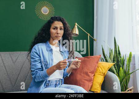 Une belle jeune femme latino-américaine est choquée par la nouvelle d'une grossesse non planifiée. Elle est assise à la maison sur le canapé, tenant un test de grossesse positif à la maison dans ses mains. Confuse, elle regarde la caméra. Banque D'Images