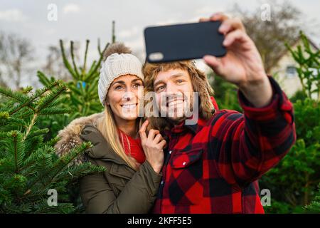 Femme et homme tenant un smartphone et souriant pour prendre un selfie sur le marché de l'arbre de noël en s'amusant Banque D'Images