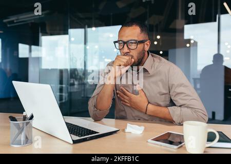 Homme d'affaires toussant au travail au bureau, homme afro-américain en lunettes et chemise malade sur le lieu de travail en utilisant un ordinateur portable au travail. Banque D'Images