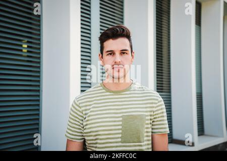 Portrait de sérieux beau beau jeune homme étudiant avec une courte coupe de cheveux debout et regardant directement la caméra isolée sur l'arrière-plan du bâtiment Banque D'Images