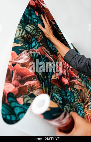 gros plan d'un homme caucasien, sur un manteau de travail gris, fixant un papier peint à motifs floraux à un mur blanc Banque D'Images