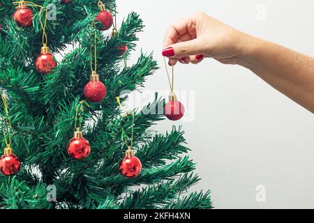 Goias, Brésil – 18 novembre 2022 : main femelle aux ongles peints en rouge, tenant une balle rouge, assemblant l'arbre de Noël. Banque D'Images