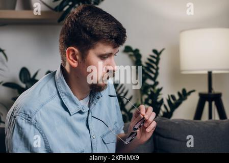 Gros plan du jeune homme caucasien réfléchi 30s travaille à l'ordinateur assis sur un canapé. Homme indépendant barbu en lunettes travaillant seul la nuit. Commande à distance Banque D'Images