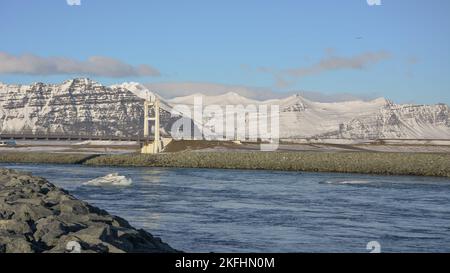Pont suspendu à voie unique au-dessus de la lagune glaciaire de Jokulsaron et de la rivière dans le sud de l'Islande. Icebergs flottant le long de la rivière. Montagnes enneigées Banque D'Images