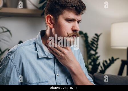 Malade jeune homme caucasien assis sur un canapé à la maison toucher le cou souffrent d'angine a mal de gorge gars malsain douleur avaler sentir inconfort coronavi Banque D'Images