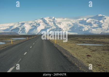 Longue route de tarmac tout droit menant au glacier islandais. Feux de route jaunes avec montagnes enneigées. Banque D'Images