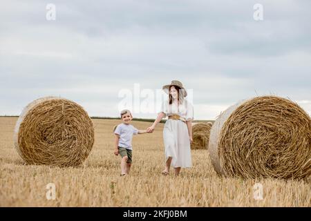 Jeune mère et jeune fils marchant près de piles de paille - balles empilées de foin laissées après la récolte, champ de ferme avec la récolte moissonnée. Banque D'Images