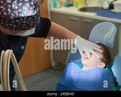 Une femme dentiste portant une casquette florale tenant la bouche d'un garçon caucasien avec la main lors d'un examen à l'hôpital dentaire Banque D'Images