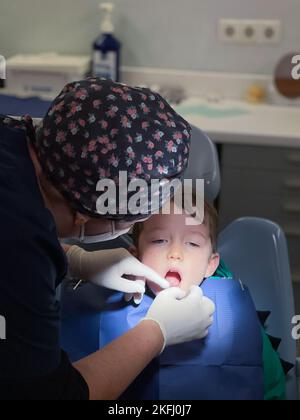 Une femme dentiste porte une casquette florale et des gants chirurgicaux pour examiner les dents d'un garçon assis sur une chaise en clinique dentaire Banque D'Images