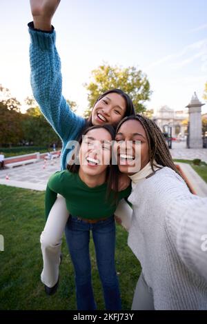 Bonne photo de trois amies différentes prenant un selfie joyeux faisant du pigeyback. Les filles s'amusent Banque D'Images