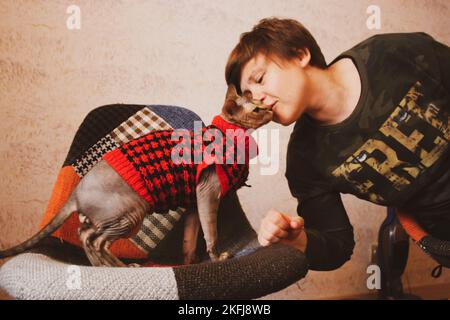 Femme embrassant le chat aimé de race canadienne de Sphynx. Mistress et chauve kitty dans un chandail rouge chaud assis sur une chaise élégante dans un intérieur moderne. Humain et pe Banque D'Images