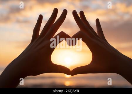 Femme mains silhouette faire coeur geste symbole sur mer coucher de soleil fond, amour émotion signe Banque D'Images