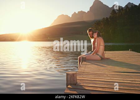 Amour au bord du lac. Jeune couple affectueux en maillots de bain assis sur un quai au coucher du soleil. Banque D'Images