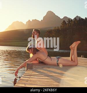 L'eau est parfaite... un jeune couple affectueux en maillots de bain assis sur un quai au coucher du soleil. Banque D'Images