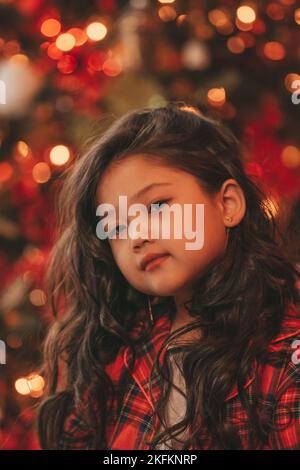 Bonne petite fille de rêve de japon regardant loin en attendant miracle de Santa posant près de l'arbre de noël. Asiatique Kid noir cheveux bouclés dans un costume rouge à carreaux fête Banque D'Images