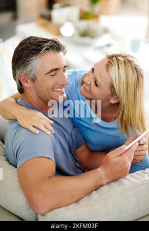 Nous avons toujours un rire ensemble. Un couple affectueux et mûr riant tout en utilisant une tablette numérique sur le canapé à la maison. Banque D'Images
