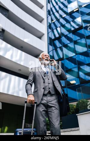 Vue à angle bas d'un professionnel souriant parlant sur un smartphone. Homme d'affaires portant un costume porte un sac pour ordinateur portable et une valise. Il est stan Banque D'Images