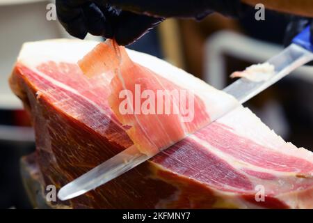 main gantée du boucher qui, avec un couteau tranchant, tranche une tranche de jambon cru Banque D'Images