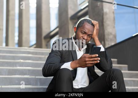 Triste homme d'affaires afro-américain lisant des nouvelles dans la dépression assis dans les escaliers devant un employé de bureau tenant un smartphone. Banque D'Images