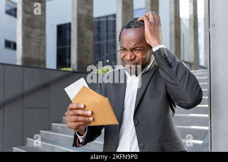 Mauvaises nouvelles, avis de faillite, homme d'affaires à l'extérieur de l'immeuble de bureaux lisant de mauvaises nouvelles dans la lettre, patron afro-américain dans le costume d'affaires déçu et triste. Banque D'Images