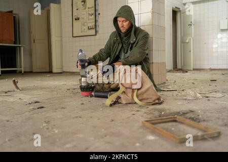 Après-apocalypse. Un jeune homme est assis sur le sol d'un bâtiment abandonné qui coule de l'eau lourde dans la tasse. Concept d'un homme qui marche dans un monde post-apocalyptique lo Banque D'Images