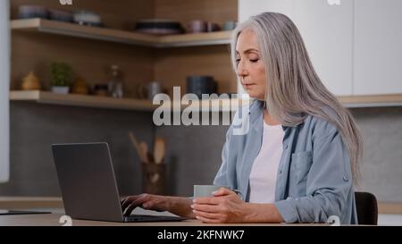 Femme d'affaires caucasienne femme au foyer senior indépendant femme mature assis à la cuisine à la maison tient une tasse de thé ou de café chaud dans les mains garde au chaud travaux sur Banque D'Images